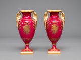 pair of red vases with gold decoration on stands against a gray background