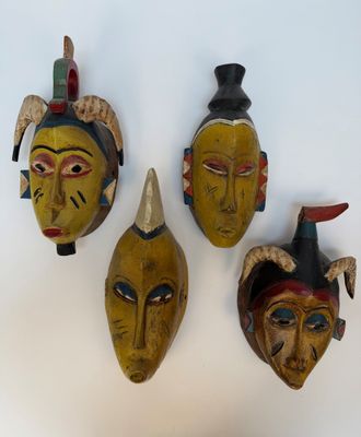 trois masques en bois ornés de cornes et de plumes, posés sur un fond blanc