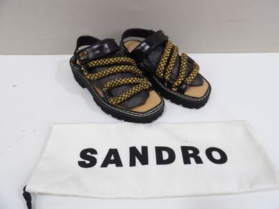 Sandales Sandro noires et jaunes à semelles noires, posées sur une table blanche