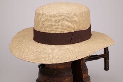 un chapeau de paille orné d'un ruban marron, posé sur un porte-chapeaux en bois