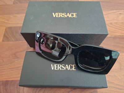 Lunettes de soleil « versa versa versa » noires et roses, avec leur étui, posées sur un sol en bois