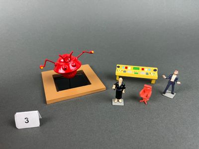 une figurine représentant un homme et une femme debout devant une table sur laquelle se trouve un objet rouge