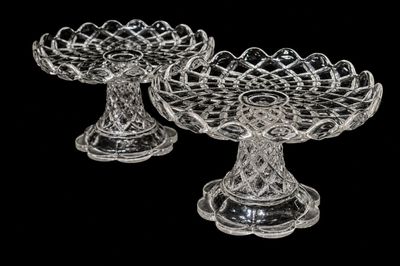 pair of clear glass footed dishes on pedestals against a black background