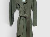 manteau en laine verte avec une ceinture, suspendu à un cintre contre un mur blanc