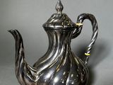 an antique silver tea pot with a twisted handle on a gray surface