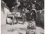 photographie en noir et blanc d'une femme en robe longue, debout dans un village