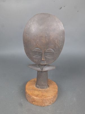 sculpture représentant un visage, posée sur un socle en bois, sur une surface grise