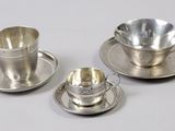 trois tasses à thé en argent avec leurs soucoupes, posées sur une surface blanche
