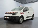 La camionnette blanche est garée dans une grande pièce ; elle porte une bande rouge sur le côté.