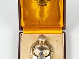 une image représentant une broche en argent dans un écrin en or, posée sur une surface blanche
