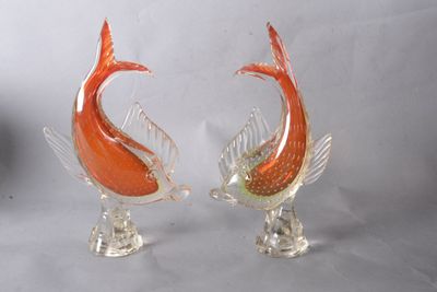 une paire de sculptures de poissons en verre orange posées sur des socles transparents, sur fond blanc