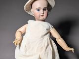 an old fashioned doll sitting on the ground wearing a white dress and a hat
