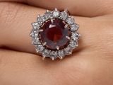 une image représentant la main d'une femme tenant une bague ornée d'une pierre rouge entourée de diamants blancs