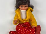 une petite poupée assise par terre, vêtue d'une veste jaune et d'un pantalon rouge