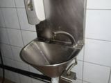 un lavabo en acier inoxydable dans des toilettes publiques, avec un distributeur de savon fixé au mur