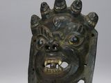 une image représentant un masque en bronze posé sur un socle métallique