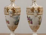 two white vases with gold trim and flowers on them sitting on a table