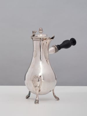 une cafetière ancienne en argent, avec une poignée noire, posée sur une table blanche