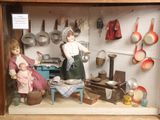 deux poupées dans une vitrine, avec des casseroles et des poêles accrochées au mur