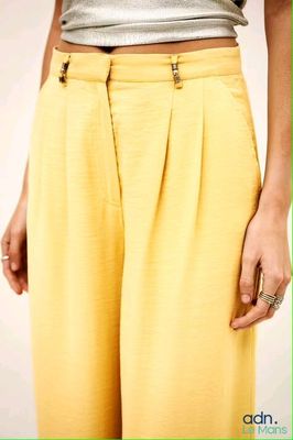 une femme vêtue d'un pantalon jaune et d'un haut argenté, debout devant un mur blanc