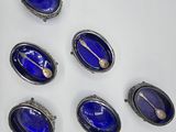 six oval blue glass brooches with spoons and spoon rest on a white surface