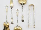 cinq cuillères et fourchettes en argent et en or, ainsi qu'un repose-cuillère, posés sur une surface blanche