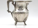 an antique silver plated pitcher on a stand on a white surface