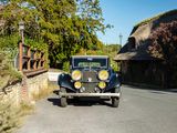 a vintage car driving down the road in front of a thatched house