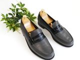 black leather loafers with gold buckles and a plant on a white surface
