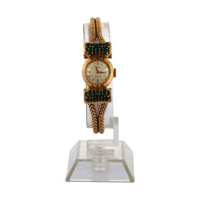 une montre en or ornée de pierres vertes, posée sur un socle transparent