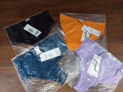 cinq t-shirts de couleurs différentes, dans des sacs en plastique, posés sur un plancher en bois