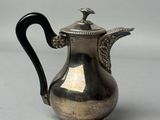 une cafetière ancienne en argent avec une poignée noire sur une surface blanche