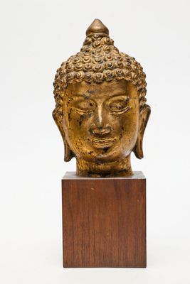 une tête de Bouddha en or posée sur un socle en bois, sur fond blanc