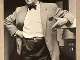 photographie en noir et blanc d'un homme en costume et nœud papillon, adossé à un mur