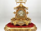 une horloge dorée posée sur un coussin rouge, sur une surface blanche