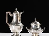 two silver teapots and a saucer on a white table