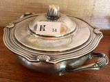 un plat ancien en argent, muni d'un couvercle et d'une poignée, posé sur une table en bois