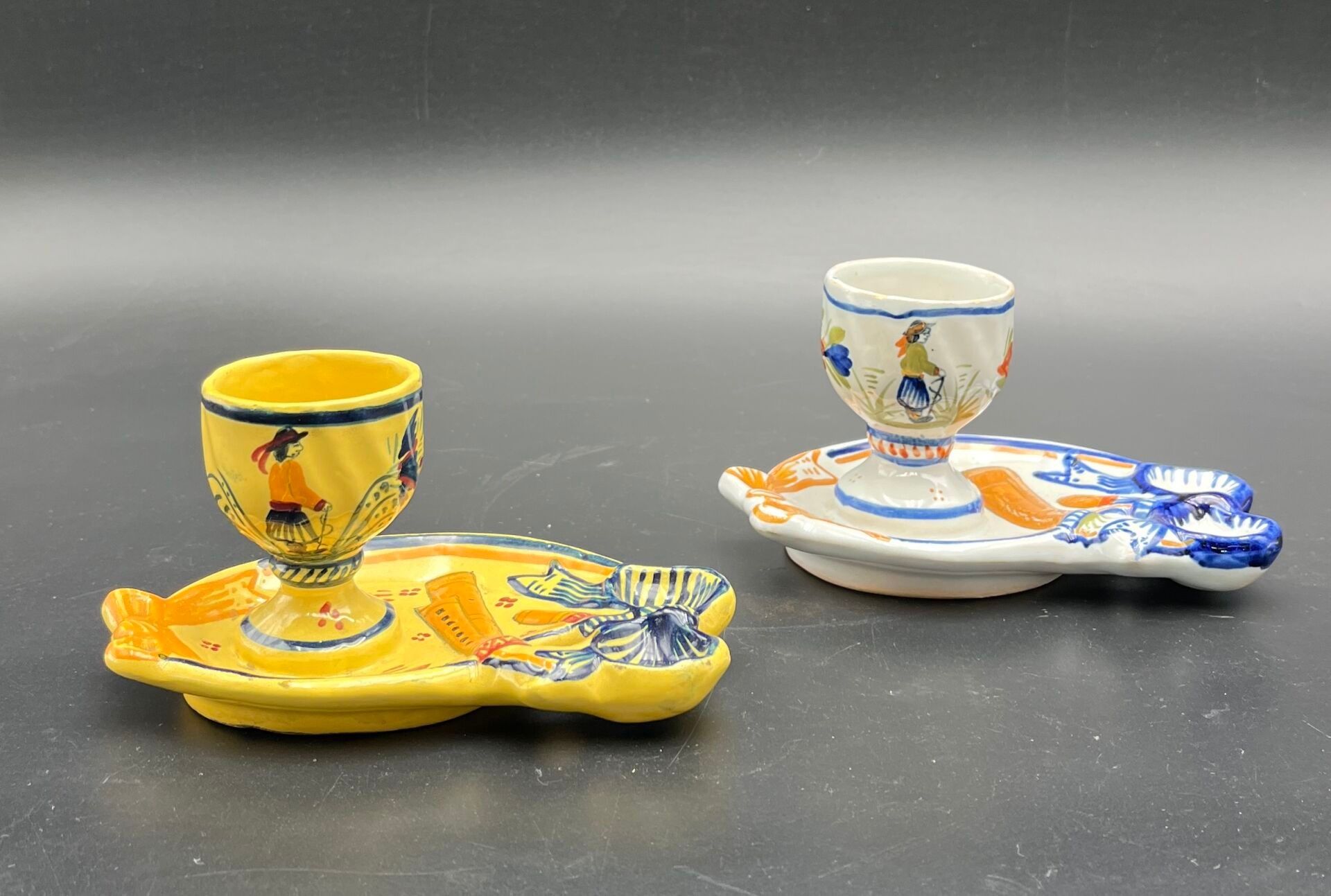 HENRIOT QUIMPER. Deux coquetiers en faïence à fond blanc et jaune à décor polychrome floral et de Bretons. 1930-1940....