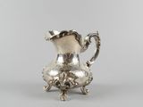 an antique silver plated pitcher sitting on a stand on a gray surface