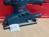 une ponceuse orbitale Bosch posée sur une table en bois, à côté d'une boîte rouge