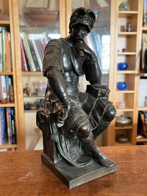 statue en bronze représentant un homme assis sur un tabouret, avec une bibliothèque en arrière-plan