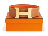 a brown and tan belt with a gold h buckle on top of an orange box