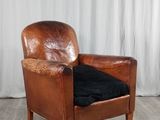 fauteuil en cuir marron avec un coussin noir, posé sur un parquet, devant un rideau blanc