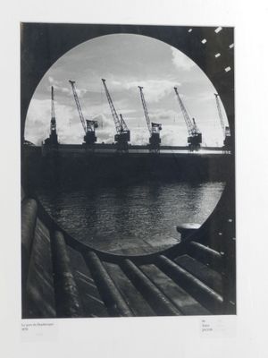 black and white photograph of a ship in the water with cranes in the background