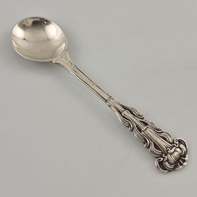 une cuillère en argent ornée d'un motif décoratif, posée sur une surface blanche