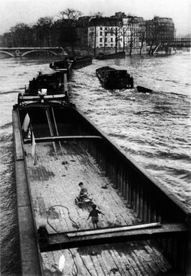 Photographie en noir et blanc d'un bateau sur une rivière, avec un pont en arrière-plan