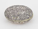 une boîte en argent richement décorée de motifs floraux, posée sur une surface blanche