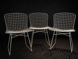 trois chaises en fil métallique blanches, alignées les unes à côté des autres dans une pièce sombre
