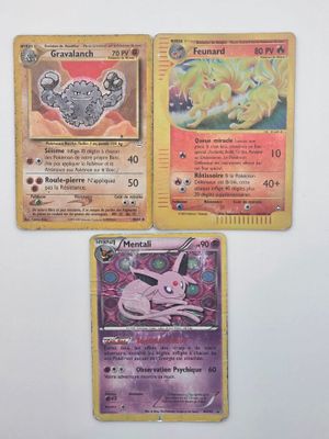 Trois cartes Pokémon sont posées sur une surface blanche ; l'une est rose et l'autre est jaune.