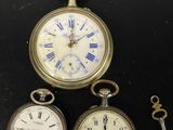 trois montres de poche anciennes et une clé posées sur un fond noir